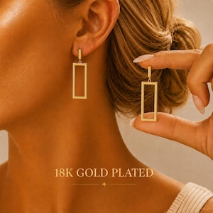 May include: A pair of 18K gold plated earrings featuring a hollow rectangular frame design. One earring hangs from an earlobe while the other is held between fingers, showcasing the metallic finish and the open geometric shape of the jewelry.