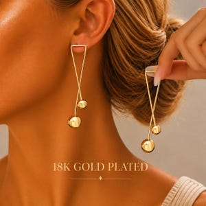 May include: A pair of 18K gold plated earrings featuring a long, thin triangular wire frame that crosses over itself. Each earring is accented with two smooth, polished gold spheres of different sizes attached to the bottom of the wire structure.