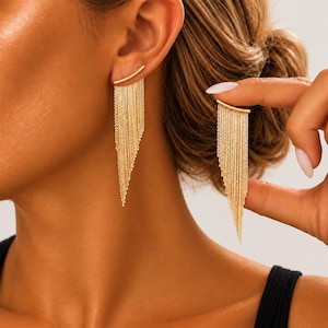 Può includere: Un paio di orecchini color oro caratterizzati da una barra curva sulla parte superiore con molteplici catenelle sottili e pendenti che creano un effetto a frangia triangolare. Gli orecchini sono color oro metallizzato e hanno una finitura lucida.