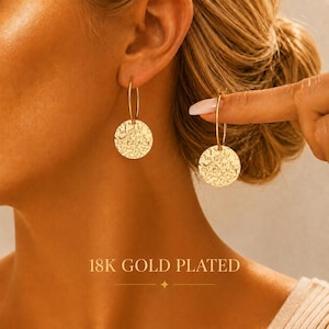 May include: A pair of 18K gold plated hoop earrings featuring a thin circular wire hoop attached to a round, textured pendant with a hammered finish. The text 18K GOLD PLATED is clearly visible at the bottom of the image in a clean, sans-serif font.