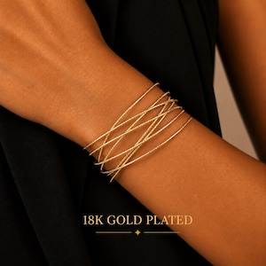 Op de afbeelding: Een set van meerdere dunne, getextureerde 18K vergulde armbanden die samen om de pols worden gedragen. De armbanden hebben een fijn, herhalend patroon op het oppervlak, wat zorgt voor een gelaagd uiterlijk tegen een donkere stof.