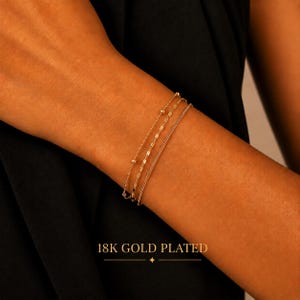 May include: A set of three 18K gold plated bracelets worn on a wrist. The set includes one delicate chain with small spherical beads, one chain with elongated oval links, and one simple fine cable chain. The text 18K GOLD PLATED is visible.