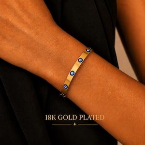 May include: A gold-toned bangle bracelet featuring a series of small, circular evil eye charms with blue, white, and black centers. The bracelet has a smooth, polished finish and is labeled with the text 18K GOLD PLATED at the bottom.