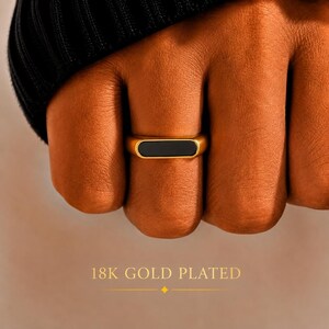 May include: A gold-toned ring featuring a smooth, rectangular black inlay centred on the band. The ring is 18K gold plated. The design consists of a polished metallic finish surrounding a flat, dark, solid-coloured rectangular centre piece.