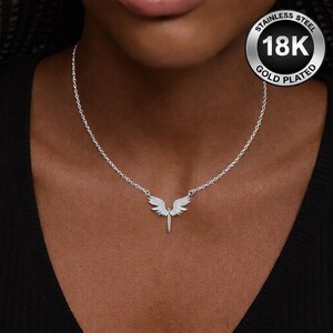 May include: A silver-colored necklace with a pendant shaped like angel wings. The wings are embellished with small, sparkling stones. The necklace is on a woman's neck. The pendant is gold plated and made of stainless steel.