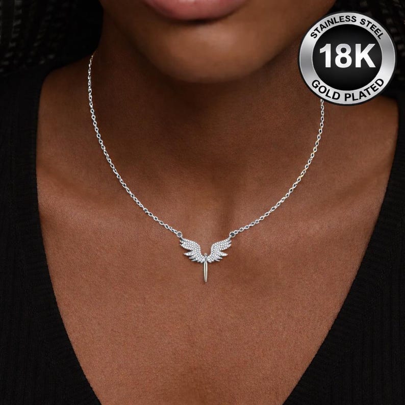 May include: A silver-colored necklace with a pendant shaped like angel wings. The wings are embellished with small, sparkling stones. The necklace is on a woman's neck. The pendant is gold plated and made of stainless steel.