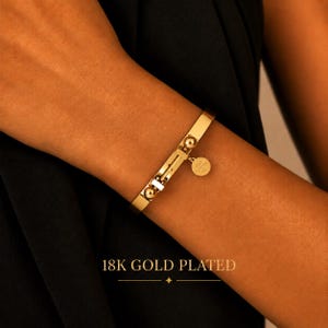 May include: An 18K gold plated bangle bracelet featuring a smooth, flat band with two spherical accents and a small circular charm. The charm is engraved with the text LOVE FOREVER 520. The bracelet has a hinged clasp mechanism for closure.