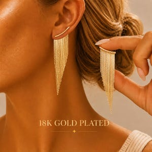 May include: A pair of 18K gold plated earrings featuring a curved bar design with multiple delicate hanging chains that create a tapered, fringe-like silhouette. The text 18K GOLD PLATED is visible at the bottom of the image.