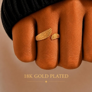 Op de afbeelding: Een 18K vergulde ring met twee gedetailleerde vleugelontwerpen die om de vinger wikkelen. Het metaal heeft een textuurafwerking met fijne lijntjes die veren nabootsen. De tekst 18K GOLD PLATED staat onderaan de afbeelding gedrukt.