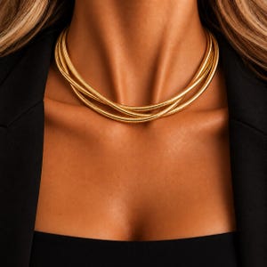 Op de afbeelding: Een goudkleurige ketting met meerdere strengen, bestaande uit verschillende gladde, flexibele slangenkettinglagen die om de nek wikkelen. De ketting heeft een gepolijste, reflecterende metalen afwerking en rust dicht bij het sleutelbeen.
