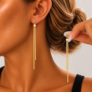 May include: A pair of gold-toned drop earrings featuring a small spherical stud connected to a square-shaped link. Two long, slender, textured chain tassels hang from each square link, creating a layered, dangling effect that extends downward.