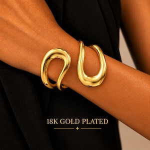 Op de afbeelding: Een 18K vergulde armband met een gladde, gepolijste afwerking en een open, vloeiende, organische vorm. Het sieraad heeft een doorlopend, gebogen ontwerp dat om de pols valt en een helder, reflecterend metalen oppervlak laat zien.