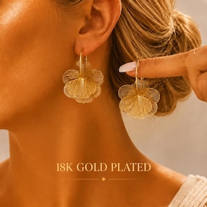 May include: A pair of 18K gold plated earrings featuring a delicate, multi-layered floral design with intricate wire-like filigree petals. The earrings are circular in shape and hang from a thin gold hoop. The text 18K GOLD PLATED is visible.