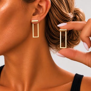 May include: A pair of gold-toned earrings featuring a minimalist rectangular open frame design. One earring is worn on the earlobe, while the other is held between fingers, showcasing the smooth, polished metal finish and geometric shape.