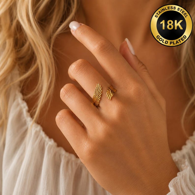 May include: A gold-plated 18K stainless steel ring with a wing design. The ring features two separate wing-shaped elements, one on each side of the finger. The ring is shown on a person's hand.
