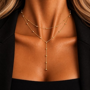 May include: A gold-toned layered necklace featuring two delicate chains adorned with small, evenly spaced spherical beads. The upper chain forms a choker style, while the lower chain extends into a vertical drop, creating a Y-shaped silhouette.