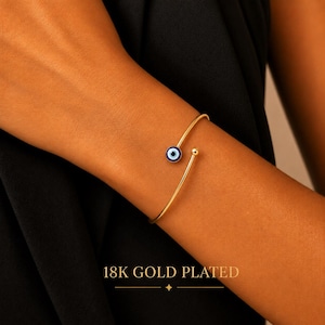 Könnte beinhalten: Ein 18K vergoldetes offenes Armband mit einem kleinen kreisförmigen Nazar-Amulett mit blauen, weißen und schwarzen Emaille-Details an einem Ende und einer glatten goldenen Kugel am anderen Ende, mit dem Text 18K GOLD PLATED darunter.