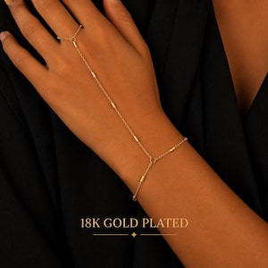 May include: An 18K gold plated hand chain featuring a delicate link design with small cylindrical accents. The jewelry connects a ring to a wrist bracelet with a thin gold chain. The text 18K GOLD PLATED is visible at the bottom of the image.