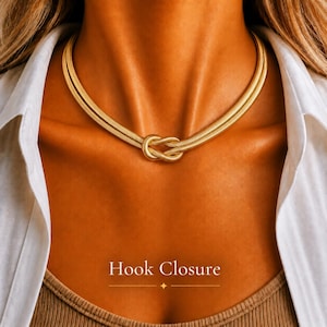 May include: A gold-toned snake chain necklace featuring a central knot design. The necklace is worn around the neck and includes a hook closure. The text Hook Closure is displayed at the bottom of the image with a small four-pointed star icon.