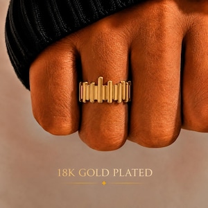 May include: A gold-plated ring featuring a series of vertical bars of varying heights arranged in a row around the band. The text 18K GOLD PLATED is displayed below the ring, indicating the material finish of the jewelry piece.