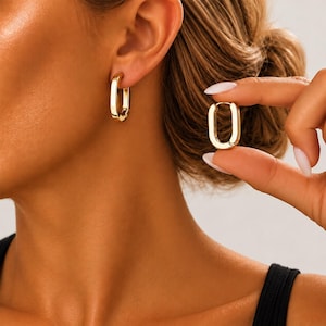 May include: A pair of gold-toned hoop earrings with an elongated, rectangular oval shape. One earring is worn in the earlobe, while the other is held between fingers to display the smooth, polished metal surface and the hinged closure mechanism.