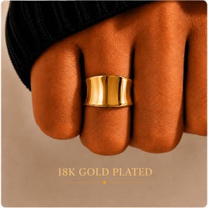May include: A wide, polished gold ring with a smooth, concave surface. The ring features a reflective, metallic finish and is labelled as 18K gold plated. The design is simple and solid, showcasing a curved band that sits across the finger.