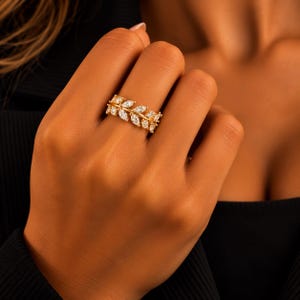 Può includere: Un anello dorato caratterizzato da una fascia con una serie di pietre trasparenti taglio marquise disposte a forma di foglia. Le pietre sono incastonate in una montatura in metallo dorato che avvolge il dito, creando un motivo botanico continuo.
