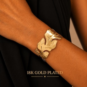May include: A gold-coloured cuff bracelet featuring a large, polished leaf-shaped design that wraps around the wrist. The surface of the metal is smooth and reflective. The text 18K GOLD PLATED is printed at the bottom of the image.