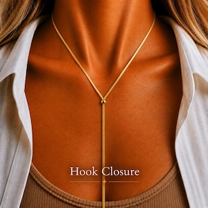 May include: A gold-toned lariat necklace featuring a flat, flexible chain that forms a Y-shape. The necklace includes a small, round bead at the centre junction where the chain splits into a vertical drop. The text Hook Closure is visible.