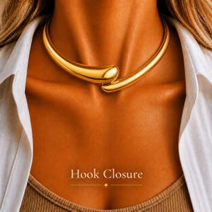 May include: A gold-toned choker necklace featuring a smooth, curved, teardrop-shaped design that overlaps at the centre. The piece has a polished, reflective surface and includes a hook closure as indicated by the text Hook Closure.