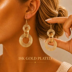 May include: A pair of 18K gold plated dangle earrings featuring a design of two interconnected circular wire coils of different sizes. The earrings hang from a thin hook wire and display a layered, concentric ring pattern in a bright gold finish.