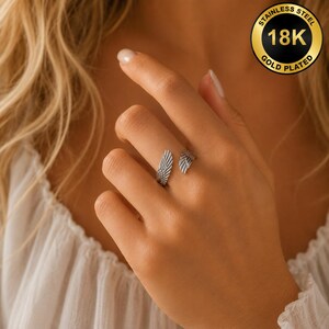 May include: A silver-colored ring with a wing design. The ring is open, with one wing on each side of the finger. The ring is made of stainless steel and is 18K gold plated.