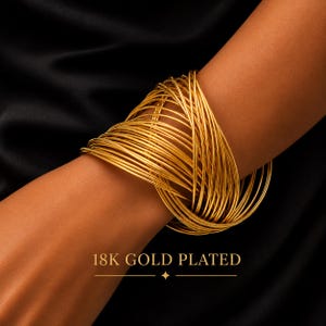 May include: A wide cuff bracelet composed of numerous thin, interlocking gold-colored wire loops. The piece features a polished finish and is labeled with the text 18K GOLD PLATED at the bottom. The design creates a dense, layered appearance.