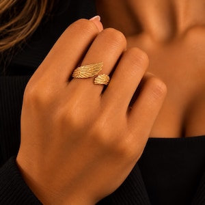 May include: A gold-toned open ring featuring two detailed wing motifs that wrap around the finger. The design showcases textured feathers on each wing, creating a layered appearance with an adjustable gap between the two ends of the band.