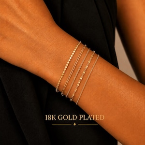May include: A set of four 18K gold plated bracelets featuring distinct chain styles, including a small disc chain, an elongated oval link chain, a textured link chain, and a simple cable chain, with the text 18K GOLD PLATED visible below.