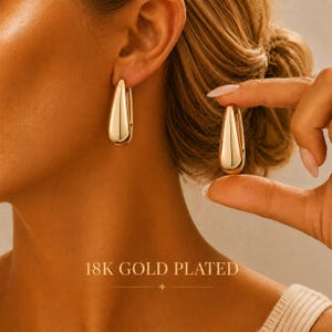 May include: A pair of 18K gold plated teardrop-shaped hoop earrings with a smooth, polished finish. One earring is worn on the earlobe, while the other is held between fingers to display the curved, solid metal design and reflective surface.