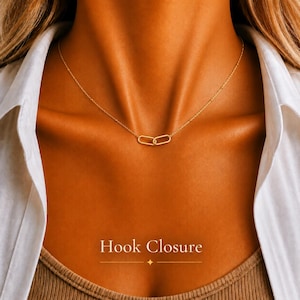 Può includere: Una collana color oro caratterizzata da una catenina sottile e due maglie ovali intrecciate al centro. Il testo Hook Closure è stampato in bianco nella parte inferiore dell'immagine, accompagnato da una piccola stella a quattro punte e una linea.