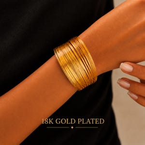 Könnte beinhalten: Ein breites Armband, bestehend aus zahlreichen dünnen, gestapelten kreisförmigen Reifen mit einer hellen, polierten 18K-Goldbeschichtung. Der Text 18K GOLD PLATED ist in einer Serifenschrift am unteren Bildrand unter dem Schmuckstück zentriert.