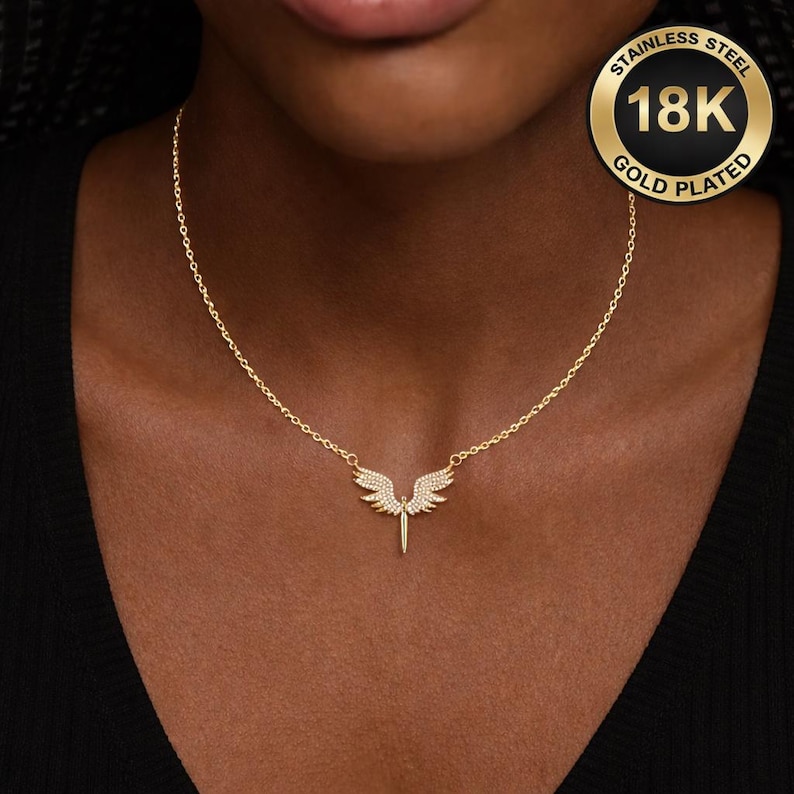 May include: Gold-plated stainless steel necklace featuring a pendant with a wing design. The pendant has a central vertical element and two wings, each adorned with small, sparkling stones. The necklace is displayed on a person's neck.