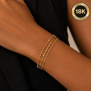 Peut inclure: Un ensemble de trois bracelets dorés présentant différents styles de chaînes, dont une chaîne à billes, une chaîne à maillons plats et une chaîne gourmette simple. Un badge circulaire affiche le texte STAINLESS STEEL 18K GOLD PLATED.