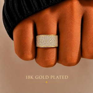 May include: A wide gold-plated band ring featuring a pavé setting with numerous small, clear, round-cut stones covering the entire outer surface. The text 18K GOLD PLATED is printed in gold lettering at the bottom of the image.