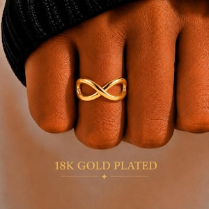 Op de afbeelding: Een 18K vergulde ring met een gepolijst oneindigheidssymbool. De ring wordt om een vinger gedragen en de tekst 18K GOLD PLATED is duidelijk zichtbaar onderaan het beeld, wat de materiaatafwerking van dit sieraad aangeeft.