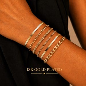 Op de afbeelding: Een set van zes 18K vergulde armbanden met diverse kettingstijlen, waaronder slangen-, stoeprand- en touwschakels. De collectie heeft een gepolijste metalen afwerking en is gestapeld om de pols gedragen. De tekst luidt 18K GOLD PLATED.