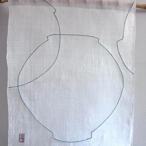 Könnte beinhalten: Ein weißer Wandbehang aus Leinen mit dunkelblaugrünen gestickten Umrissen von zwei Vasen. Der Stoff hängt an einer Holzstange. Ein kleines rotes gesticktes Quadrat befindet sich in der unteren linken Ecke des rechteckigen Textilstücks.