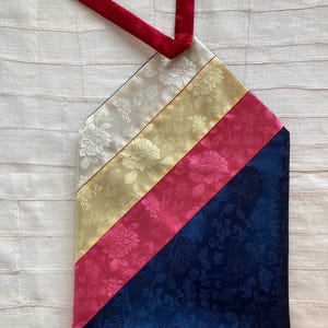 May include: A patterned pouch with a red strap. The pouch is divided into diagonal sections of white, yellow, pink, and navy blue fabric, each with a floral design. A red strap extends from the top, for carrying.