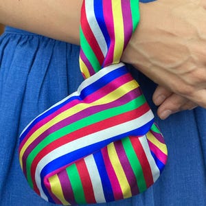 Puede incluir: Un bolso de mano a rayas de colores con un asa anudada. El bolso presenta bandas vibrantes de rojo, amarillo, verde, azul, morado y blanco. El bolso lo sostiene una persona que lleva un vestido azul.