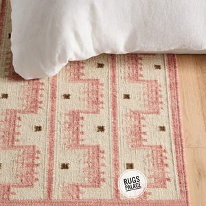 May include: A rectangular rug with a geometric pattern in shades of pink and beige, with a white pillow on top. The rug has a border and features a repeating design with small brown square accents. The rug has the text "Rugs Palace".
