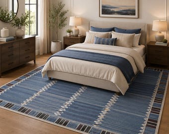 Op de afbeelding: Een groot rechthoekig vloerkleed met een blauwe basis, een herhalend wit geometrisch ruitpatroon en een donkerbruine en zwarte gestreepte rand. Het kleed ligt op een houten vloer onder een bed met beige beddengoed en blauwe accenten.