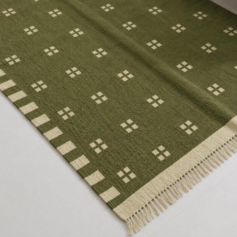 May include: An olive green woven rug featuring a repeating pattern of small, cream-colored squares arranged in a grid. The rug is finished with a wide, cream-colored border containing a striped pattern and short, cream-colored fringe along the edge.