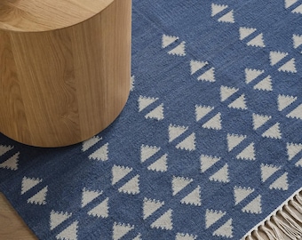 Könnte beinhalten: Blauer Teppich mit einem geometrischen Muster aus weißen Dreiecken. Der Teppich hat an einer Kante Fransen und steht neben einem runden Beistelltisch aus Holz. Das Design verleiht jedem Wohnraum eine moderne Note.